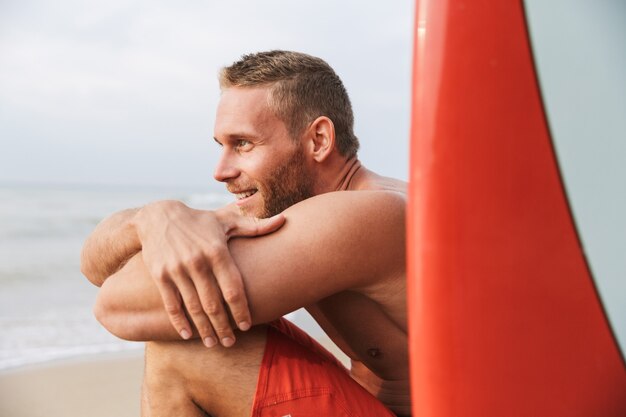 Schöner lächelnder junger Mann, der sich an einem Strand mit Surfbrett entspannt