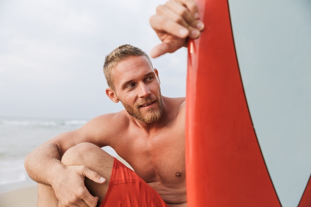 Schöner lächelnder junger Mann, der sich an einem Strand mit Surfbrett entspannt