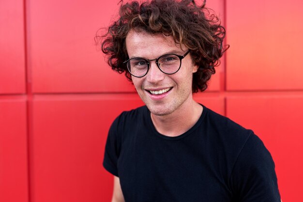 Schöner lächelnder Mann, der für Werbung an der roten Wand im Freien posiert und schwarzes T-Shirt und runde Brille mit Kopierplatz für Ihren Informationstext trägt Menschen Emotionen
