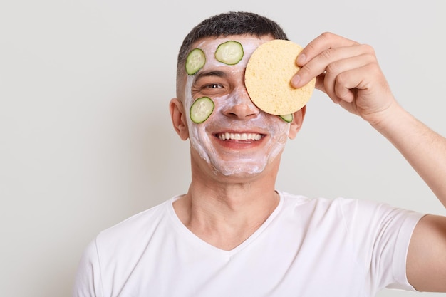 Schöner lächelnder Mann mit weißem T-Shirt, der Hautpflegeverfahren mit maskierten Gurkenscheiben durchführt, die in die Kamera schauen und das Auge mit einem Schwamm bedecken, der isoliert vor grauem Hintergrund posiert