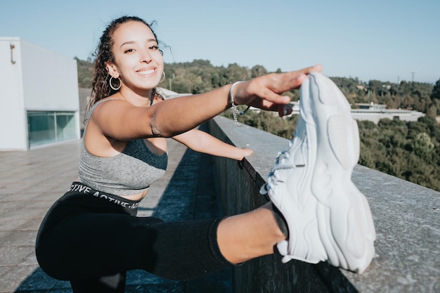 Schöner Mädchenathlet, afrikanische gebräunte Haut. Lächelt in die Kamera, während die Muskeln aufgewärmt werden, bevor sie urbanes Yoga-Training machen. Beingelenke Gelenkgymnastik. Sichere Bewegung an der frischen Luft. Leggings und Top.