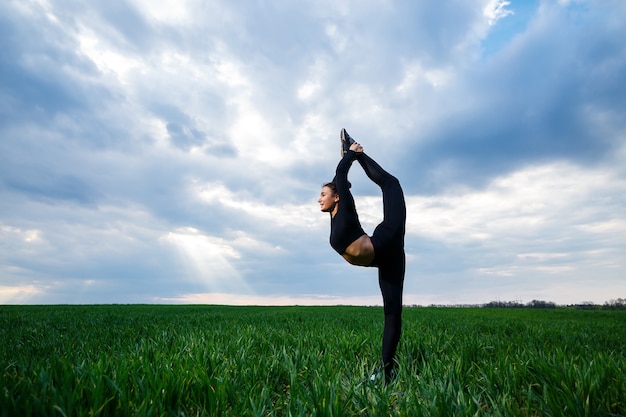 Schöner Mädchenturner auf dem grünen Gras macht Yoga. Eine schöne junge Frau auf einem grünen Rasen führt akrobatische Elemente durch. flexible Turnerin in Schwarz macht einen Handstand im Split