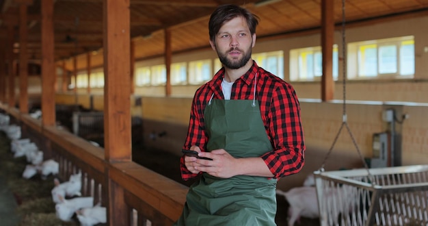 Foto schöner männlicher bauer, der im ziegenstall sitzt und eine sms-nachricht auf dem smartphone schreibt mann bauer tippt und scrollt auf dem handy moderner viehzuchtbetrieb