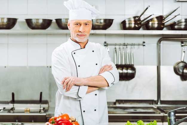 Schöner männlicher Koch in Uniform mit verschränkten Armen, der in der Restaurantküche in die Kamera blickt
