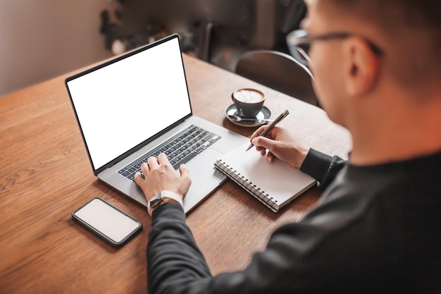 Schöner Mann, der am Arbeitsplatz am Laptop arbeitet Geschäftsmann, der Informationen auf dem Computer am Arbeitstisch mit Kaffeetelefon und Notizblock eingibt