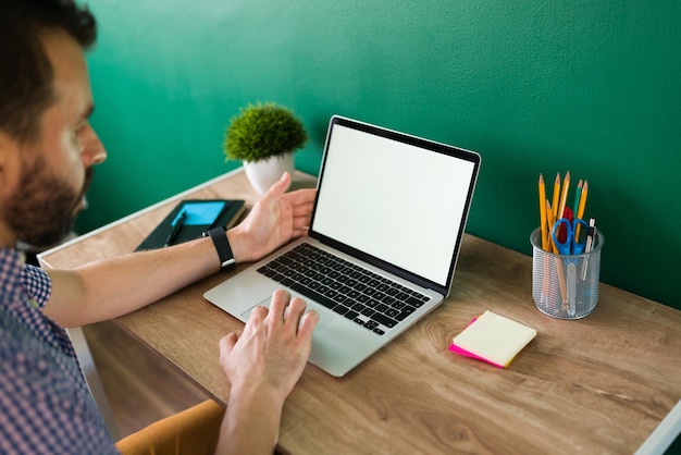 Schöner Mann, der an seinem Schreibtisch sitzt, während er auf einem Laptop mit leerem Bildschirm und Kopierbereich tippt