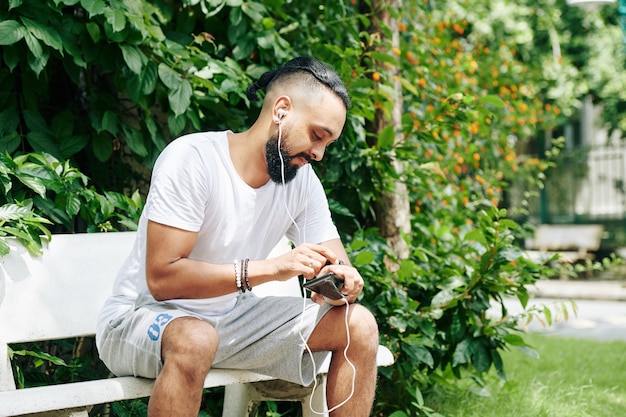 Schöner Mann, der auf einer Bank im Park sitzt, Musik auf dem Smartphone hört und die Smartwatch überprüft