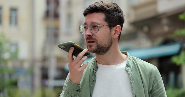Schöner Mann, der beim Gehen auf der Straße der Stadt eine Audionachricht auf dem Smartphone aufzeichnet Der Mann verwendet das Smartphone, um Sprachnachrichten im Freien zu senden Online-Sprachanruf