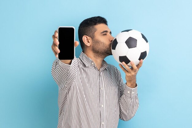 Schöner Mann, der Fußball küsst und Handy mit leerem schwarzem Display zeigt