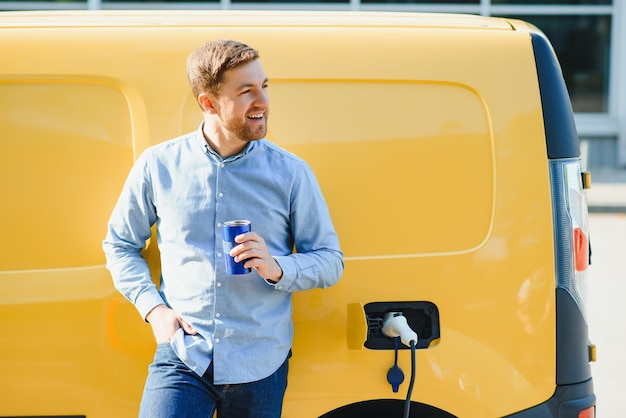 Schöner Mann, der Kaffee trinkt, während er Elektroauto auflädt