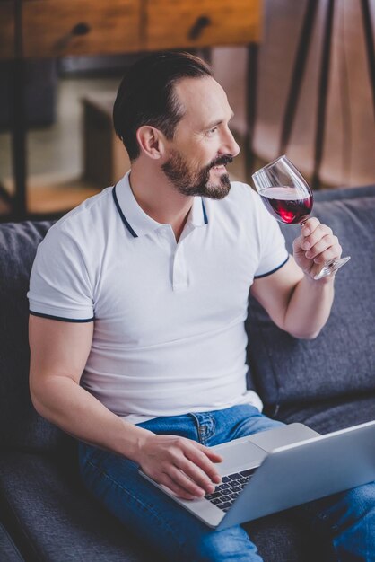 Schöner Mann, der zu Hause Laptop benutzt und Rotwein trinkt