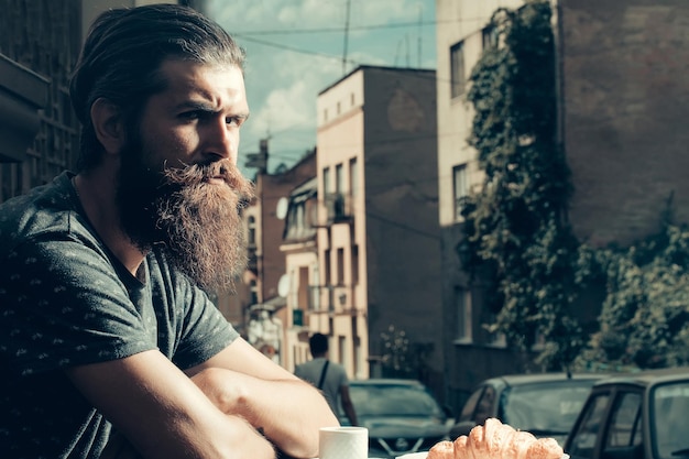 Schöner Mann Hipster mit langem Bart und Schnurrbart mit Croissant und Kaffeetasse im Café im Freien an