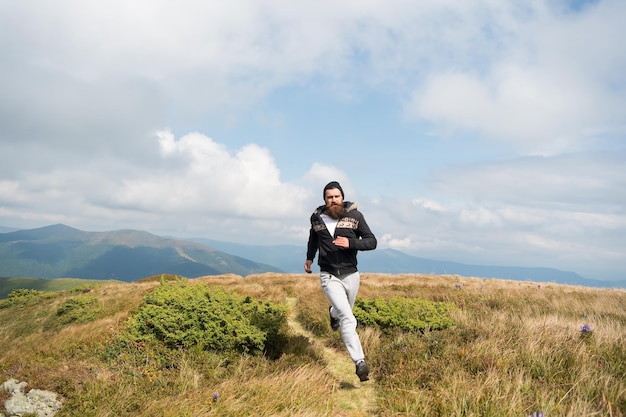 Schöner Mann Hipster oder Kerl Läufer mit Bart und Schnurrbart in Hut laufen sonnig im Freien auf Berggipfel mit grünem Gras vor bewölktem Himmel auf natürlichem Hintergrund