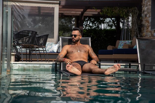 Schöner Mann im Pool auf einem Stuhl sitzend, Mann mit großem Bart, Badehose und Tattoos