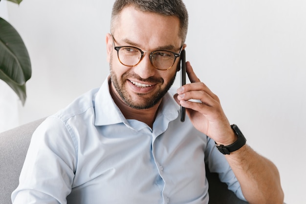 schöner Mann im weißen Hemd, der lächelt und auf schwarzem Smartphone über Geschäft spricht, während er im Büro arbeitet