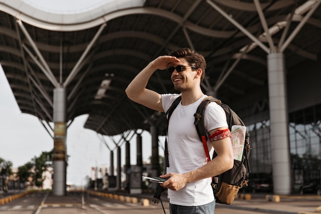 Schöner Mann in Jeans und weißem T-Shirt hält Pass und Rucksack Cooler brünetter Kerl mit Sonnenbrille posiert in der Nähe des Flughafens