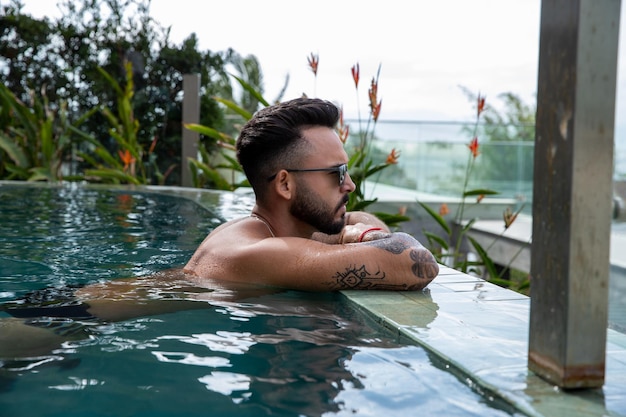 Schöner Mann mit Brille, Bart und Tattoos, der sich an einen Infinity-Pool lehnt, Mann am Fuß