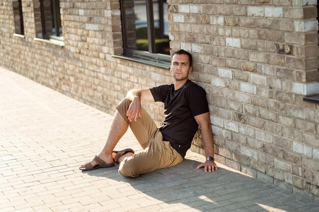 Schöner Mann mit kurzen Borsten in einem schwarzen T-Shirt auf dem Hintergrund einer Backsteinmauer, die auf dem Boden sitzt