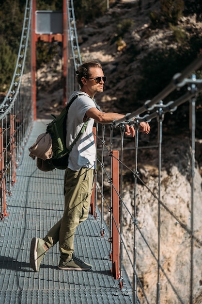 Schöner Mann mit Rucksack, der an einem guten sonnigen Tag auf der Hängebrücke über dem See steht