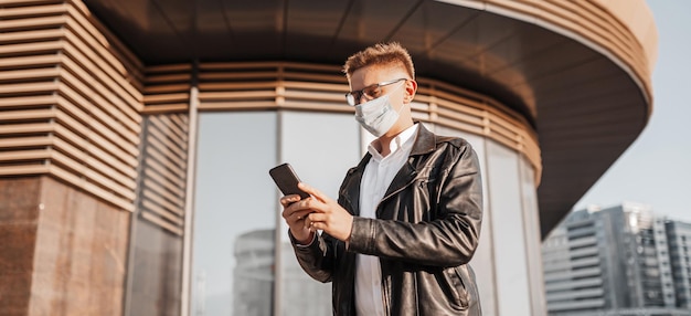 Schöner Mann mit Schutzmaske im Gesicht mit Brille mit Smartphone auf der Straße einer Großstadt. Geschäftsmann, der am Telefon auf städtischem Hintergrund spricht