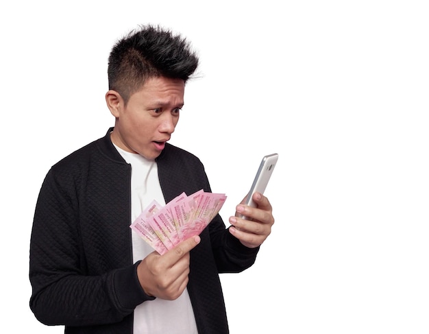 Schöner Mann mit überraschtem Gesichtsausdruck, der auf den mobilen Bildschirm blickt und indonesisches Geld in der Hand hält