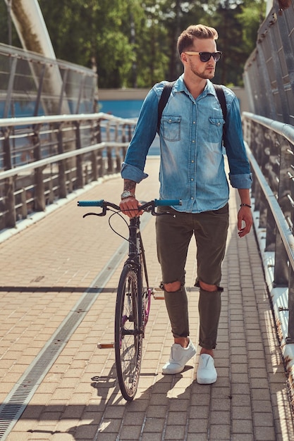 Schöner modischer Hipster mit stylischem Haarschnitt in Sonnenbrille, der mit dem Fahrrad im Freien spazieren geht.