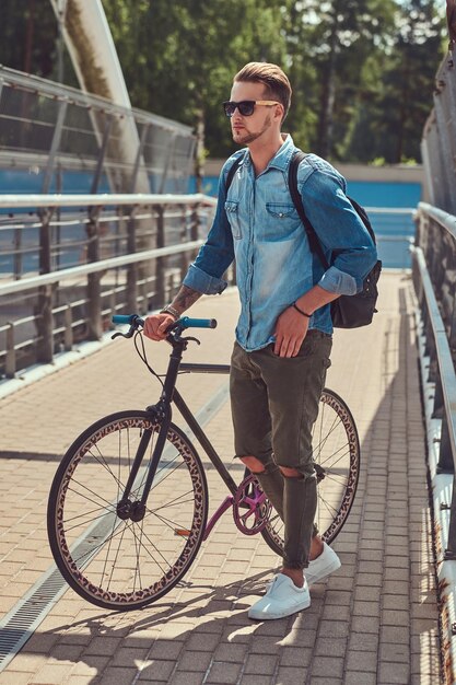 Schöner modischer Hipster mit stylischem Haarschnitt in Sonnenbrille, der mit dem Fahrrad im Freien spazieren geht.