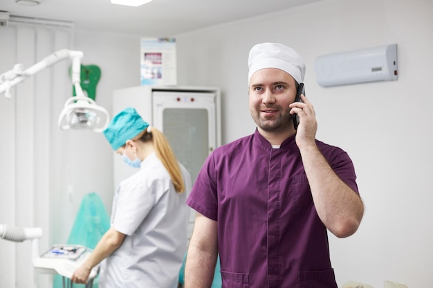 Schöner positiver männlicher Zahnarzt, der lächelt, während er in einer Zahnklinik mit dem Handy telefoniert