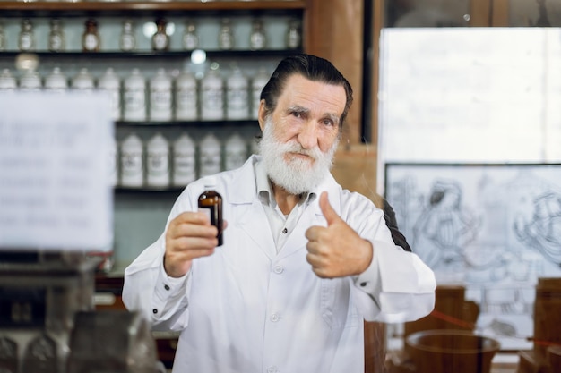 Foto schöner reifer bärtiger apotheker, der in einer alten vintage-drogerie posiert und eine flasche mit medikamenten in der hand hält, die lächelnd in die kamera schaut und den daumen nach oben zeigt