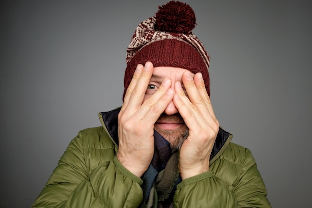 Schöner reifer Mann, der die Augen mit den Händen bedeckt, während er grauen Hintergrund in warmer Winterkleidung steht