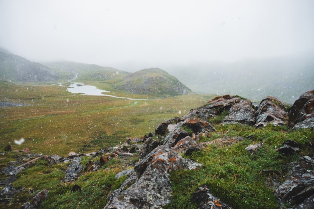 Schöner Schneefall in den Bergen. Atmosphärische grüne Alpenlandschaft mit großen Schneeflocken. Kleiner See, Wasserläufe und felsiger Hügel mit scharfen, schroffen Steinen bei Schneefall. Schneeflocken im Hochland