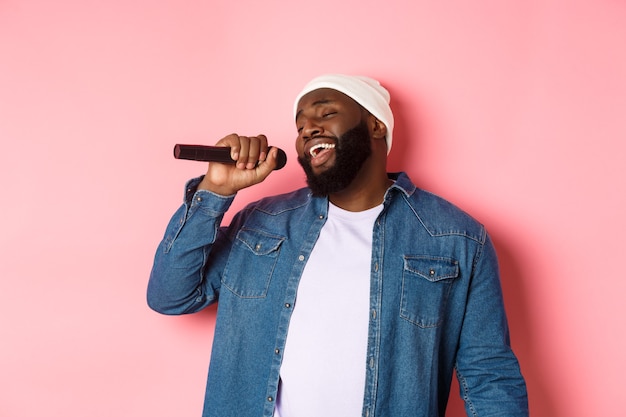 Schöner schwarzer Mann in Mütze und Jeanshemd, der Karaoke singt, Mikrofon hält und über rosa Hintergrund steht