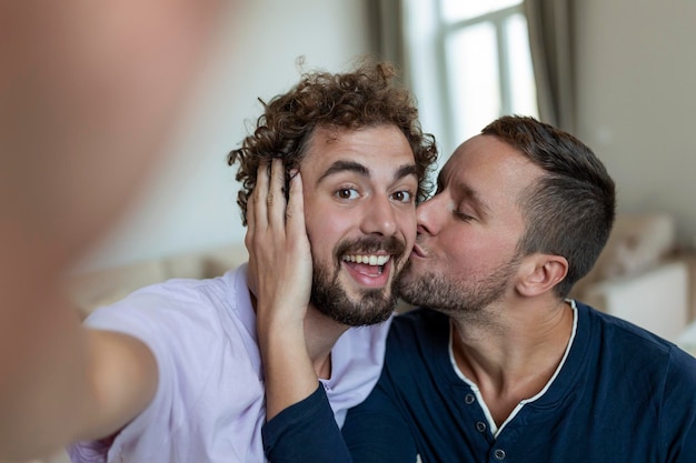 Schöner schwuler Mann, der zu Hause zusammen sitzt und sich mit dem Handy selbst macht Schwuler Mann mit seinem Partner, der sitzt, indem er ein Selbstporträt mit dem Smartphone macht