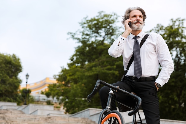 Schöner selbstbewusster bärtiger Mann mit Anzug, Aktentasche, der mit dem Fahrrad im Freien spazieren geht, mit dem Handy spricht