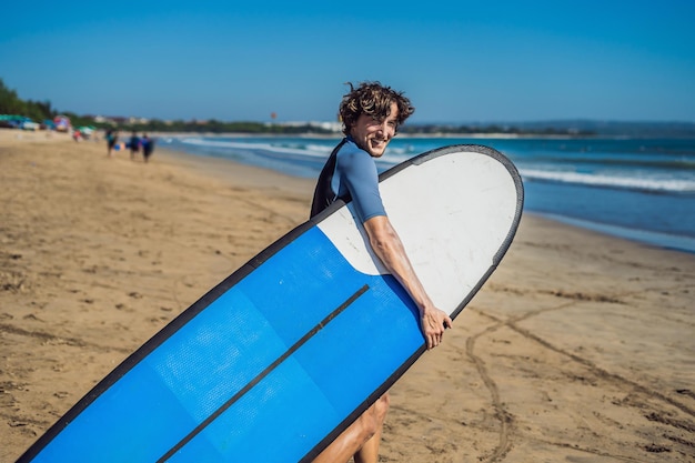 Schöner sportlicher junger Surfer, der mit seinem Surfbrett unter dem Arm in seinem Neoprenanzug an einem tropischen Sandstrand posiert