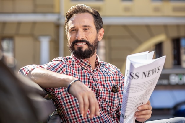 Schöner Start in den Tag. Fröhlicher hübscher Mann, der eine Zeitung hält, während er auf der Bank sitzt