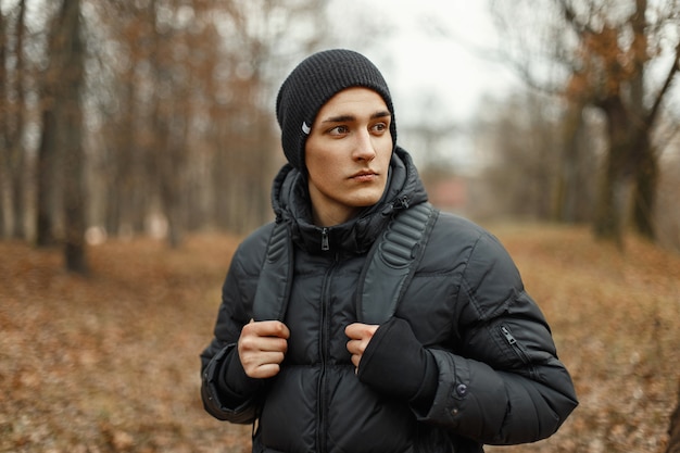 Schöner stilvoller junger Mann in der Winterjacke und im Hut mit Rucksack
