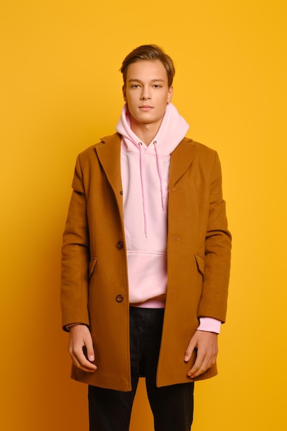 Schöner, stylischer Mann in braunem Trenchcoat und rosa Sweatshot mit Kapuze, der über gelbem Studiohintergrund posiert