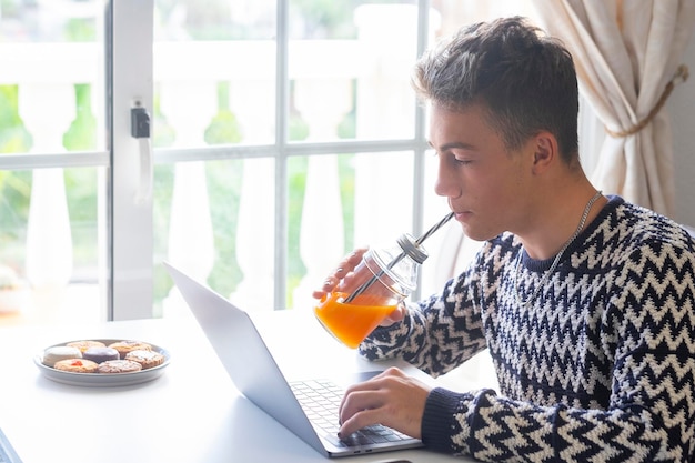 Schöner Teenager, der zu Hause einen Orangensaft mit Laptop vor dem Fenster trinkt Millenial-Generation, die Technologie und Soziales genießt