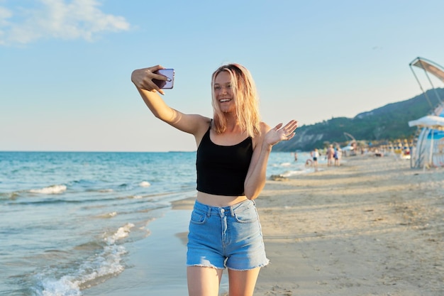 Schöner Teenager mit Smartphone, der bei Sonnenuntergang am Meeresstrand spazieren geht und ein Selfie macht