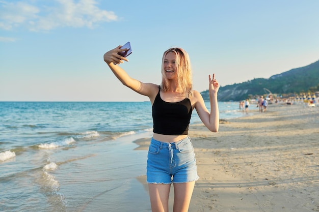 Schöner Teenager mit Smartphone, der bei Sonnenuntergang am Meeresstrand spazieren geht und ein Selfie macht