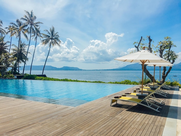 Schöner tropischer Swimmingpool im Hotel oder Resort mit Sonnenschirm, Kokosnussbaum, Sonnenliegen, Palmen mit Infinity-Pool-Blick auf das Meer und die Berge im Hintergrund