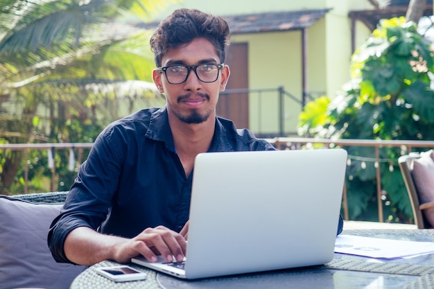 Schöner und erfolgreicher indischer Mann in einem stilvollen, gut gekleideten Freiberufler, der mit einem Laptop am Strand arbeitetFreiberufler und FernarbeiterGeschäftsmann Student in einem Sommercafé am Ufer des Indischen Ozeans