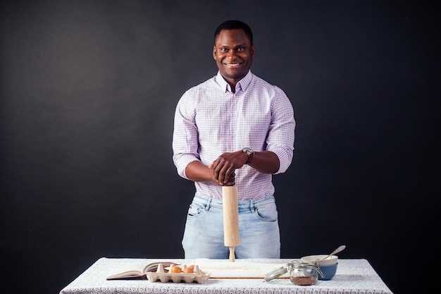 Schöner und junger afroafrikanischer Mann, der hausgemachte Kuchen zubereitet American Pie aus frischen Teighänden, die mit Mehl verschmutzt sind, auf dem Tisch sind Eier, Nudelholz und Rezeptbuch auf schwarzem Hintergrund im Studio