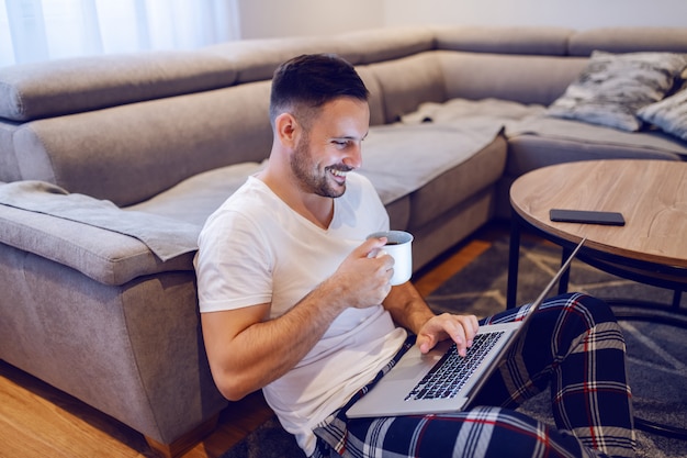 Schöner unrasierter lächelnder kaukasischer Mann, der auf Boden im Wohnzimmer sitzt, im Internet surft und am Morgen Kaffee trinkt.