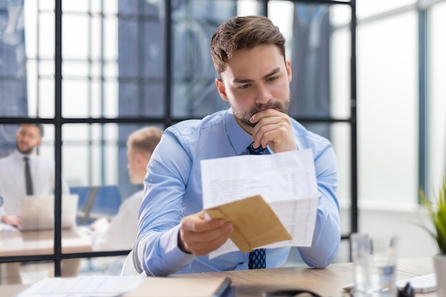 Schöner Unternehmer, der einen Brief aus dem Umschlag auf einem Schreibtisch im Büro liest, mit Kollegen im Hintergrund