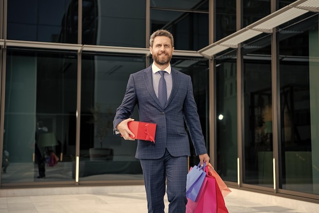 Schöner Unternehmer im Anzug mit Einkaufstüten und Geschenken spazieren vor dem Bürojubiläum