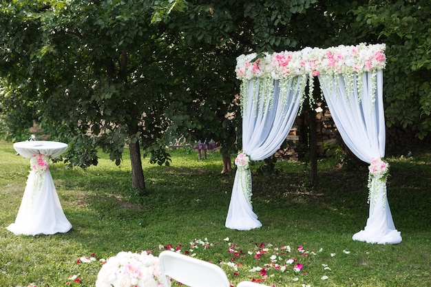 Schöner weißer Bogen mit Blumenschmuck für die Hochzeitszeremonie mit Stühlen für Gäste auf grünem Hintergrund mit Bäumen