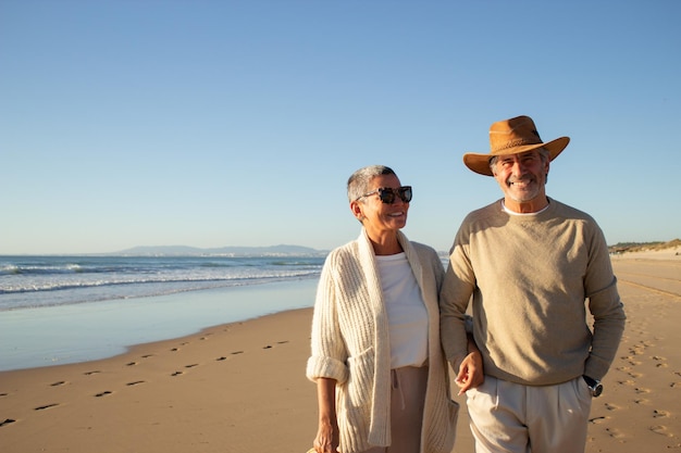 Schönes älteres Paar, das an der Küste steht, während es gemeinsam die Urlaubszeit genießt. Schöner grauhaariger Mann mit Cowboyhut und Dame mit Sonnenbrille, die glücklich lächelt. Ruhestand, Freizeitkonzept