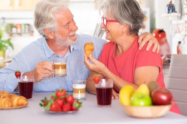 Schönes älteres Paar, das einander in die Augen schaut, während es zu Hause mit Croissant und Cappuccino frühstückt Senioren entspannt und glücklich genießen gesunde Ernährung